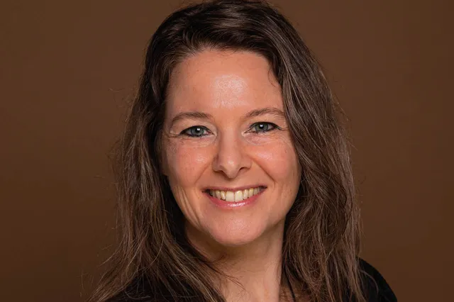 Anke Verhagen staat voor een bruine achtergrond. Ze lacht in de camera en heeft haar bruine haar los hangen.