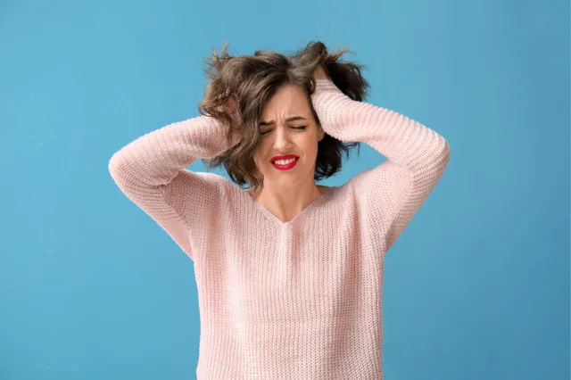 Vrouw met lichtroze trui aan en handen in het haar. Kijkt moeilijk door stress.