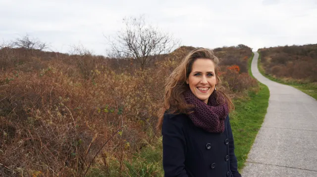 Anne staat op de foto in de natuur, ze lacht naar de camera en draagt een dikke jas en sjaal. De lucht is grauw.