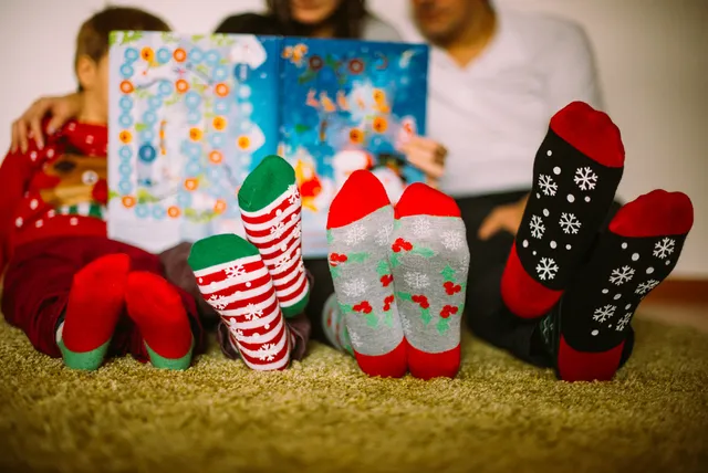 Familie zit op de grond met een boek voor hun gezichten. Ze dragen kersttruien en op de voorgrond zie je vier paar voeten met kerstsokken aan/.