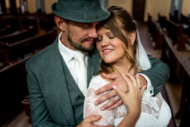 Man met groen trouwpak en hoedje omarmt zijn kersverse vrouw van achteren. De vrouw heeft een sluier en trouwjurk aan, lacht met ogen dicht naar haar man.
