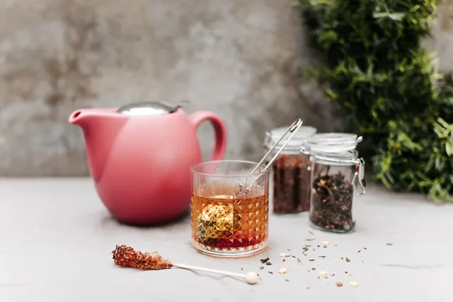 Eens wat anders op je kersttafel: vega(n) kerst-high tea. Er staat een rode theepot, met een kopje thee en twee glazen potjes verse kruidenthee naast. Achter zie je een deel van een kerstkrans.