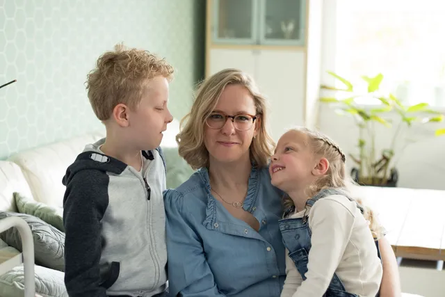 Op de afbeelding staat Jacqueline met haar zoon en dochter. Jacqueline zit in het midden en heeft halflang, blond haar en een bril op. Haar kinderen zijn allebei blond.