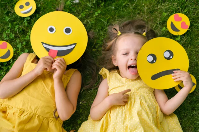 Twee kinderen in het gras met emoji's voor hun gezicht: ze leer je je kind gezond omgaan met emoties.