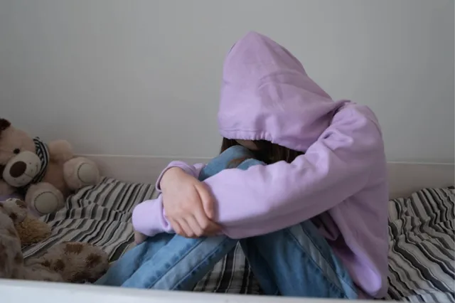 Een jonge vrouw zit op bed met haar hoofd naar beneden en een capuchon over haar hoofd, ze lijkt verdrietig.