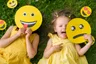 Twee kinderen in het gras met emoji's voor hun gezicht: ze leer je je kind gezond omgaan met emoties